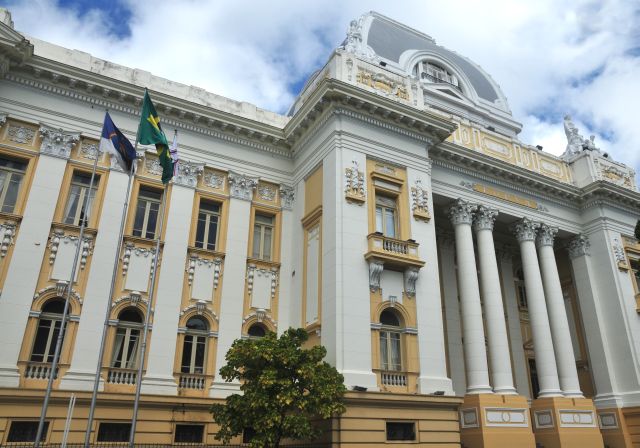 Fachada do Palácio da Justiça de Pernambuco, com as bandeiras de Pernambuco, do Brasil e do TJPE em primeiro plano e céu azul com poucas nuvens