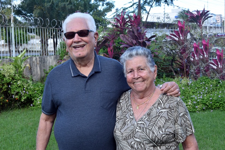 Casal Arnaldo e Sônia no jardim da casa. Ao fundo flores, plantas, e a grama da casa 