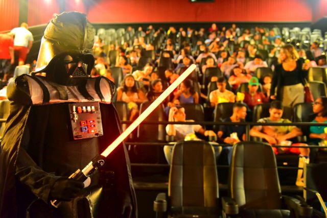 Crianças, ao fundo da sala de cinema, observam personagem Darth Vader com sabre de luz