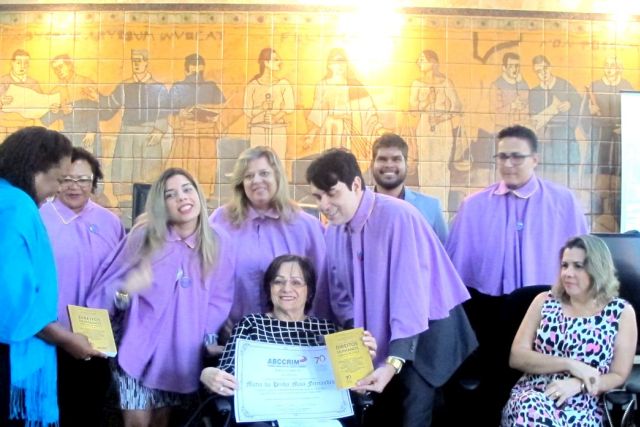 Maria da Penha sentada na cadeira de rodas rodeada por estudantes vestidos com a beca roxa e certificados na mão
