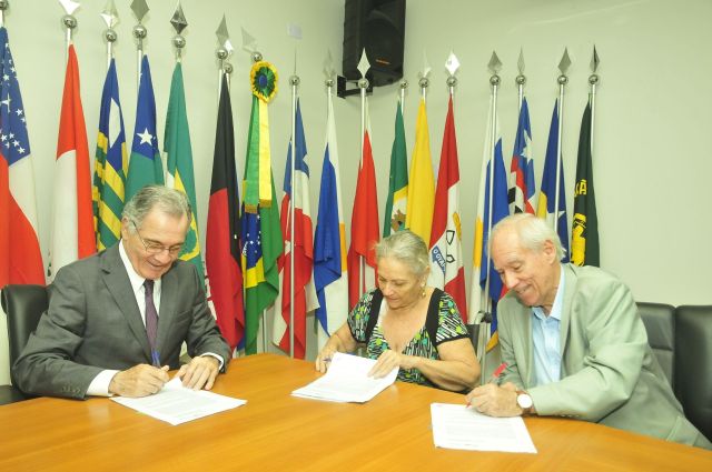 Representantes do TJPE e da Fundaj assinam acordo simultaneamente em três cópias. Documentos sobre mesa de madeira; ao fundo, bandeiras do Brasil e de outros estados brasileiros