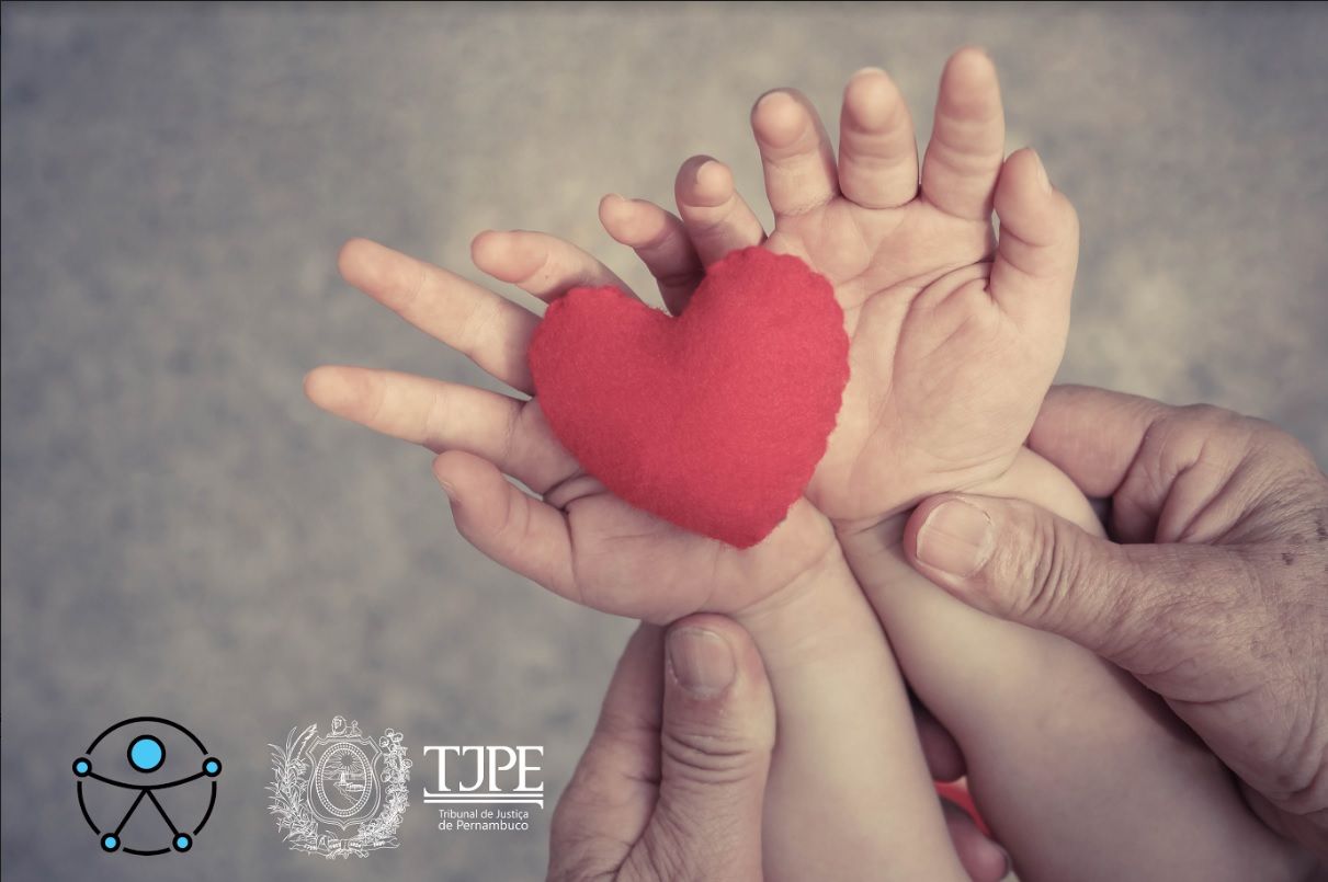 Foto das mãos de um bebê segurando coração de pelúcia apoiadas nas mãos de um adulto