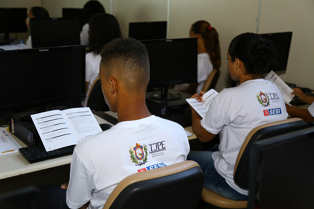 Estudantes, em frente a computadores, leem cartilha do curso de informática