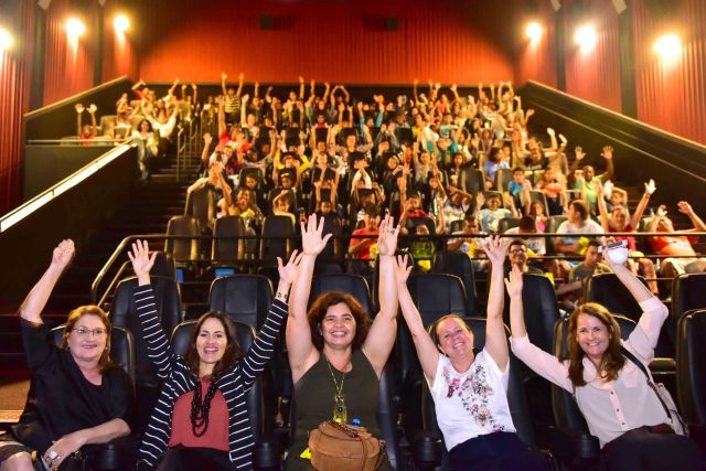 Público no cinema, sentado nas poltronas e com as mãos levantadas
