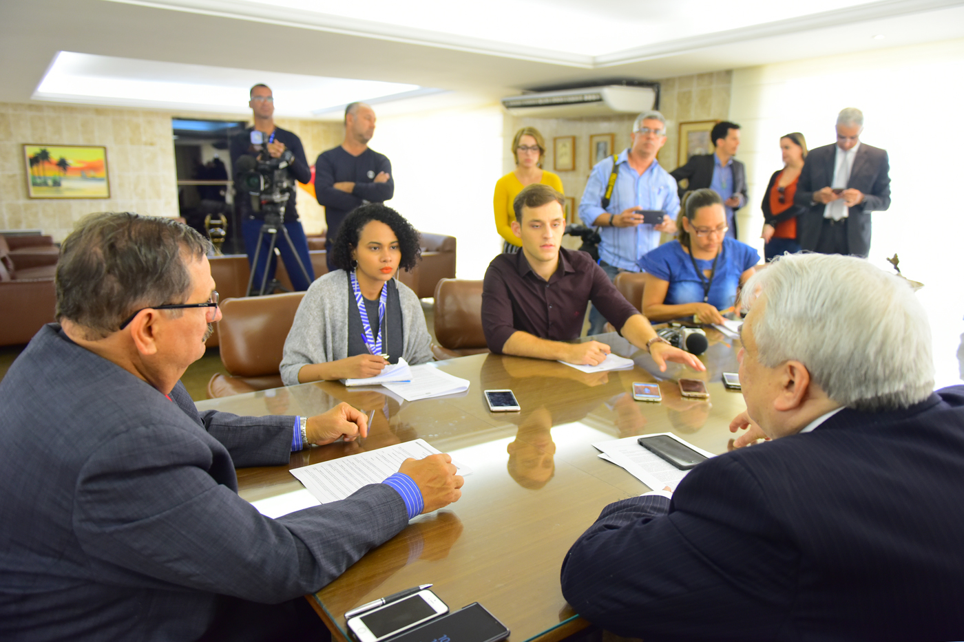 Imagem de mesa de reunião com desembargadores e jornalistas e ao fundo fotógrafos e cinegrafistas acompanhando o evento