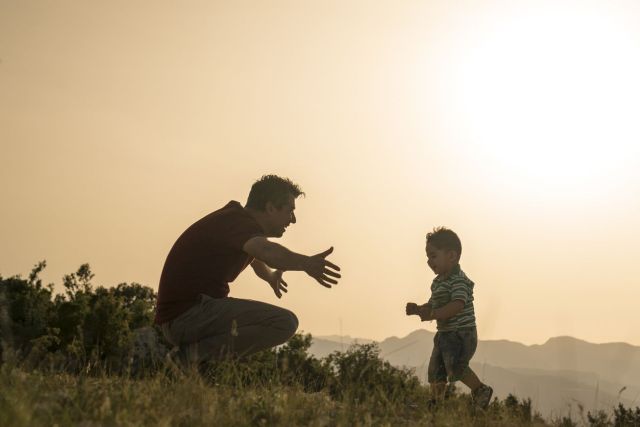Em uma paisagem iluminada pelo por-do-sol um pai abre os braços para abraçar seu filho 