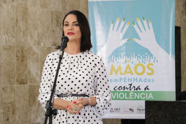 A modelo Luiza Brunet discursa em pé em frente a banner oficial do programa Mãos emPENHAdas