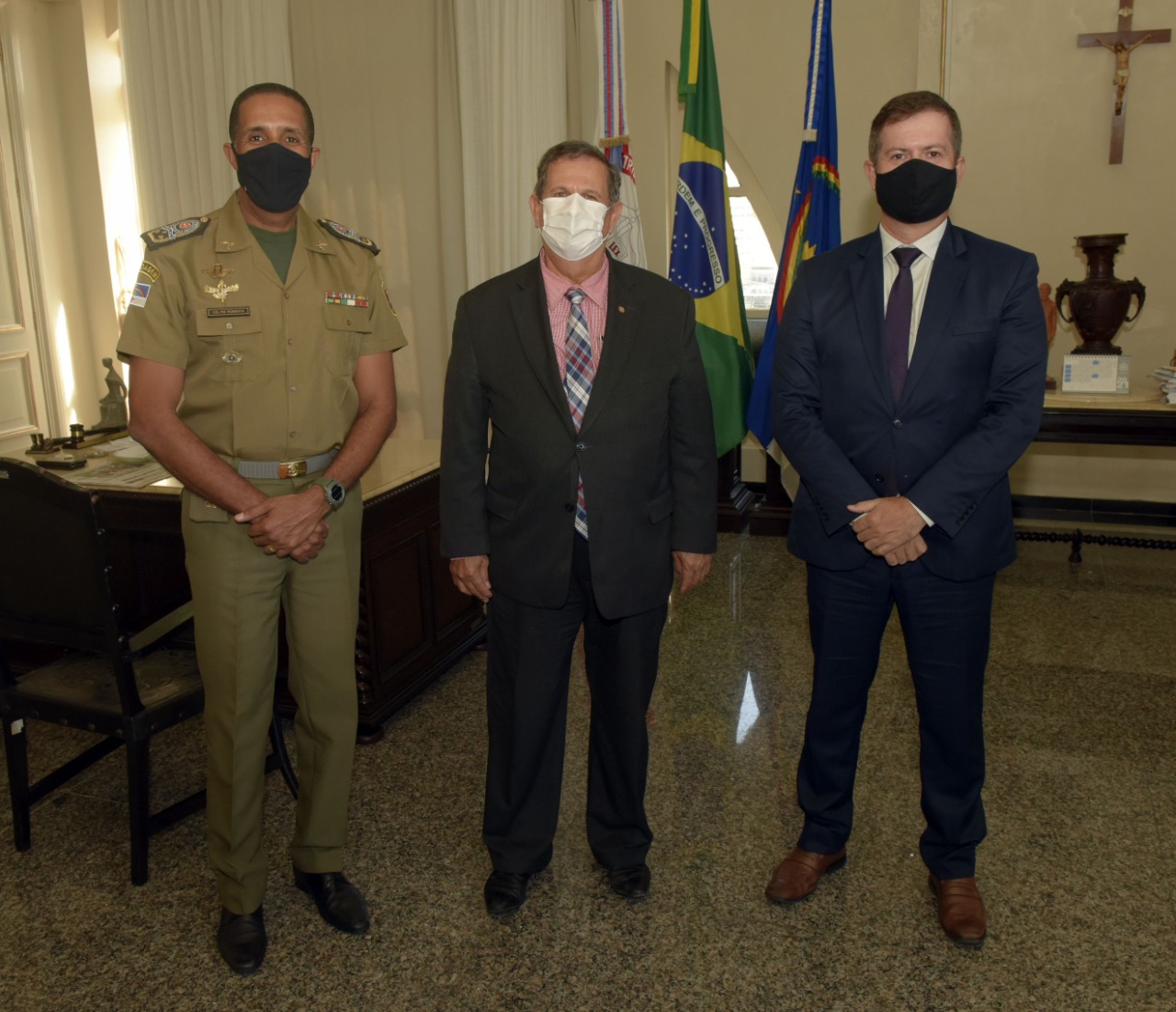 Três autoridades em um gabinete posam em pé para foto. Um deles está com fardamento militar, e os outros dois estão com trajes formais.