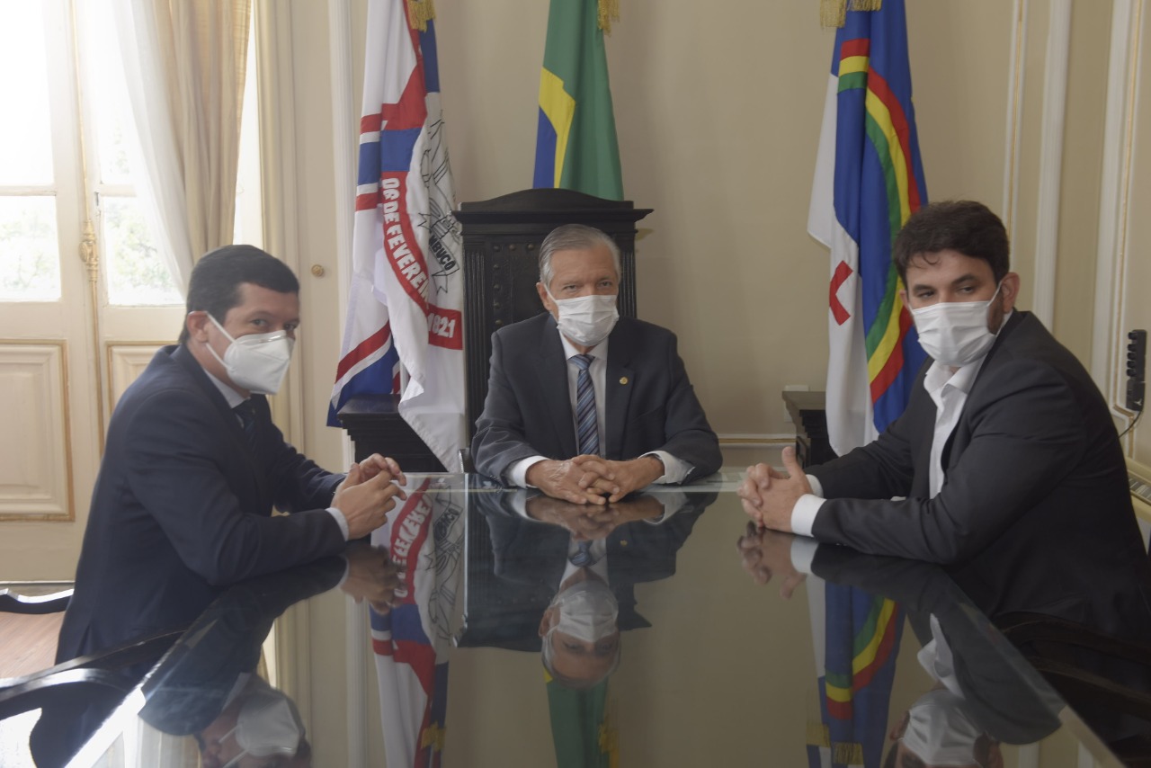 Três homens usando máscara, conversam sentados à mesa. Ao fundo, as bandeiras do TJPE, de Pernambuco e do Brasil