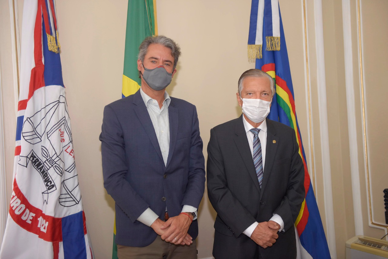 Dois homens de pé, lado a lado, posam olhando para a foto. Ao fundo, as bandeiras do Brasil, de Pernambuco e do TJPE