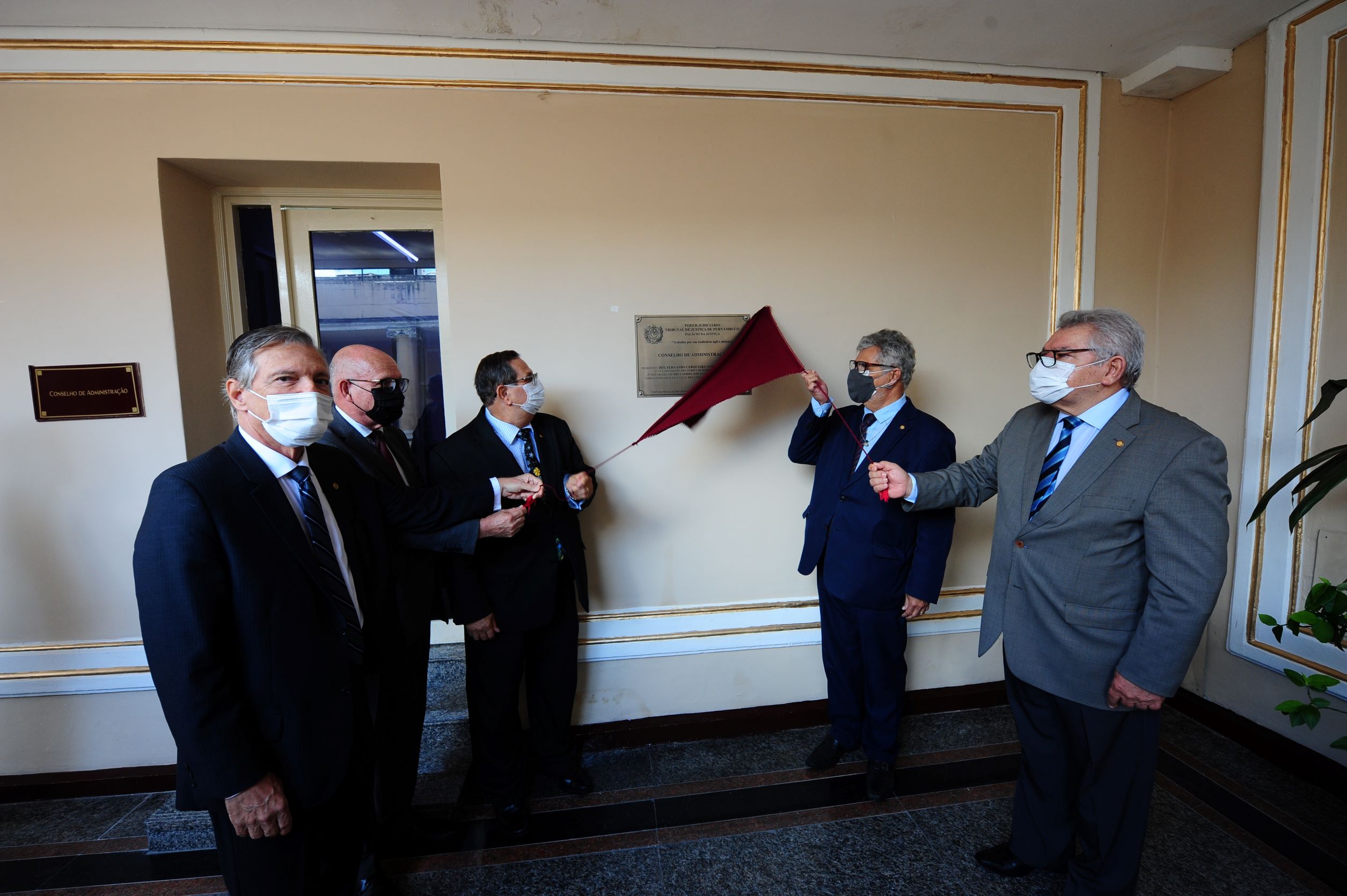 Imagem mostra cinco homens puxando um pano cor de vinho que cobre uma placa de inauguração