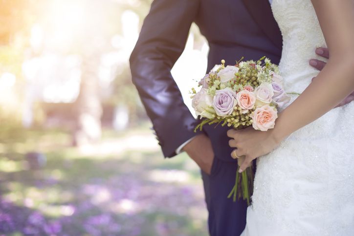 Imagem mostra noivo e noiva abraçados com destaque para a mão dela segurando o buquê