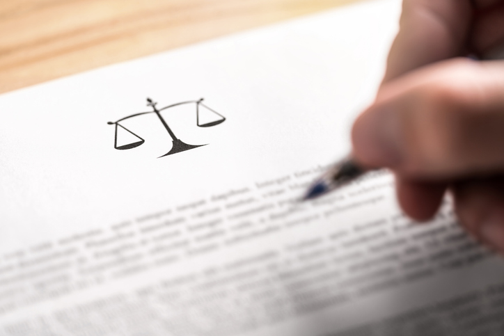 Imagem de mão segurando uma caneta diante de um texto jurídico. No topo da página, há um ícone de uma balança como sinônimo de equilíbrio, palavra que sempre é associada a Jusitça.