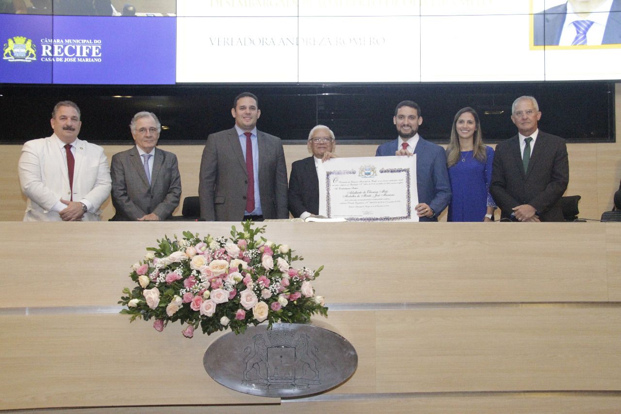 Pessoas aparecem de pé, lado a lado, em plenário. Ao centro, o homenageado segura um diploma.