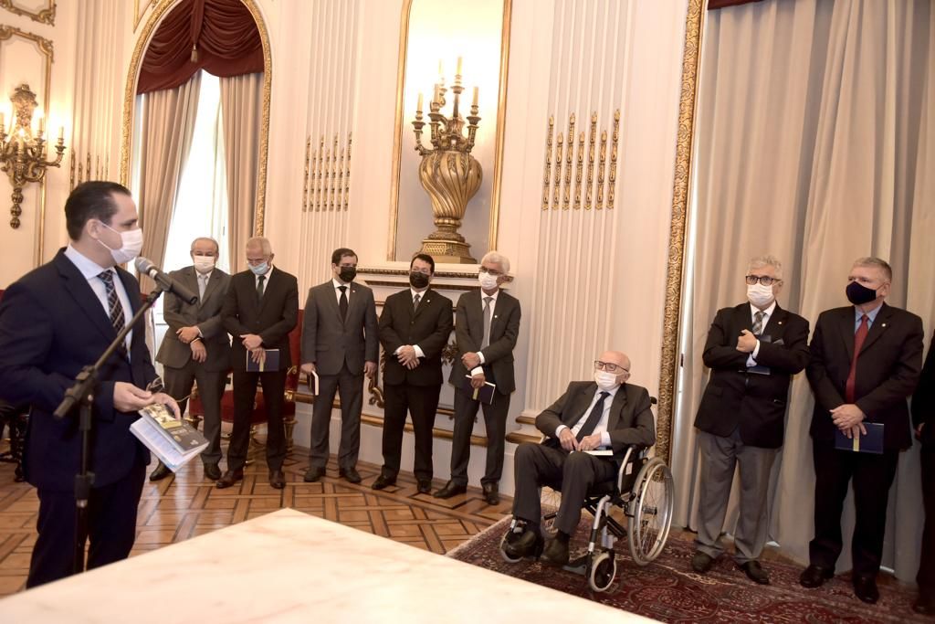 À esquerda, homem fala ao microfone para demais pessoas em uma sala. Entre elas está o homenageado sentado em uma cadeira de rodas