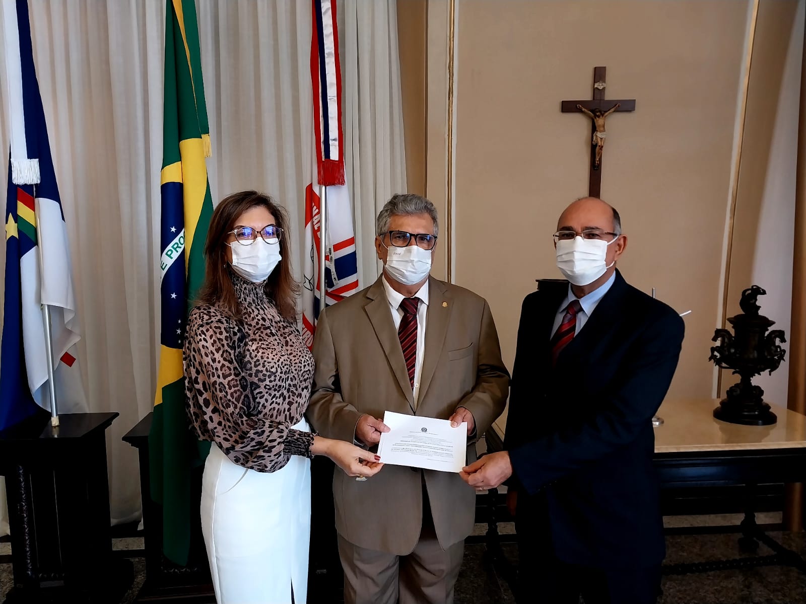 Foto mostra uma mulher e dois homens de pé, segurando um papel, olhando para a câmera. Ao fundo aparecem as bandeiras de Pernambuco, do Brasil e do TJPE