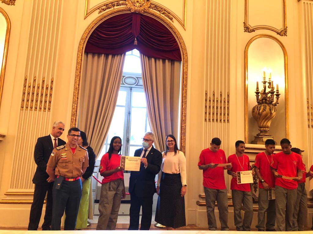 Foto mostra adolescente recebendo certificado. Ao lado estão outros jovens com seus certificados e pessoas sorrindo para a foto.