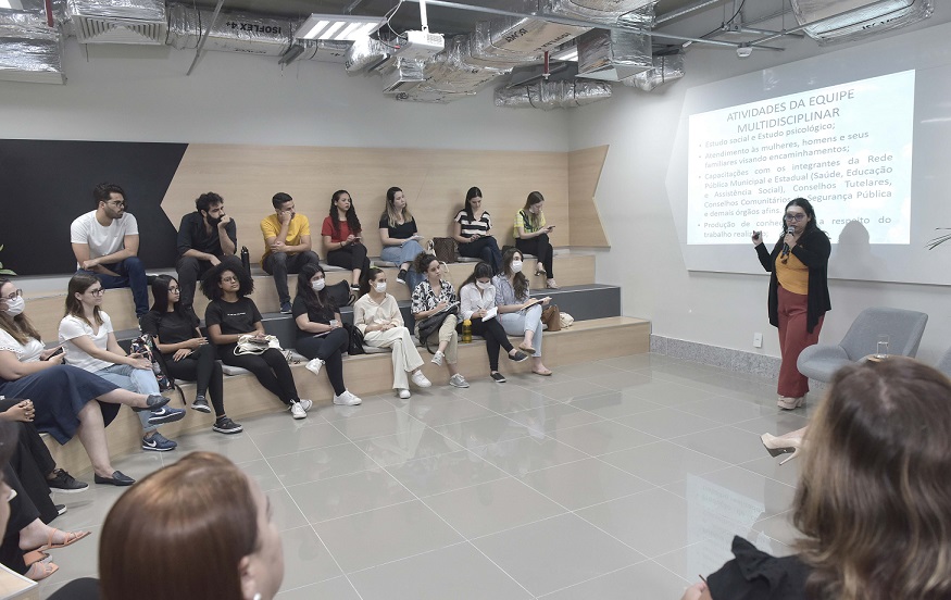 Foto mostra alunos sentados em semicírculo, olhando para mulher que fala ao microfone à frente de um quadro branco, com tela projetada.