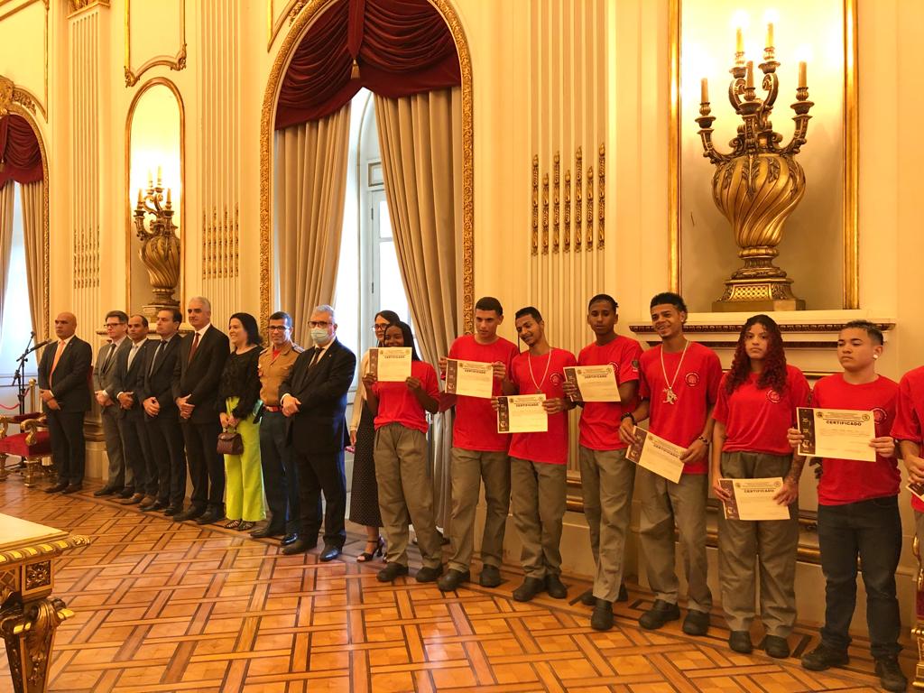 Foto mostra pessoas de pé, em um salão decorado com cortinas e candelabros. Jovens com camisas vermelhas seguram um certificado.