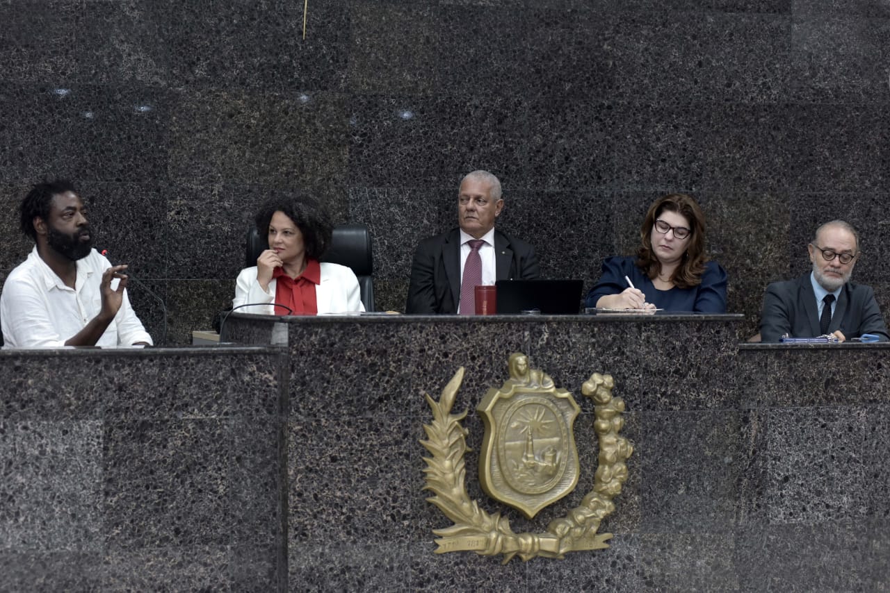 Foto mostra três homens e duas mulheres sentados lado a lado em uma plenária. À frente da bancada aparece o brasão dourado do Judiciário.