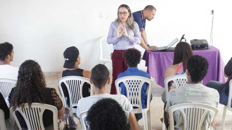 Foto da sala onde ocorreu a palestra. A assistente social Camila Almeida está em pé falando para uma plateia de adolescentes.