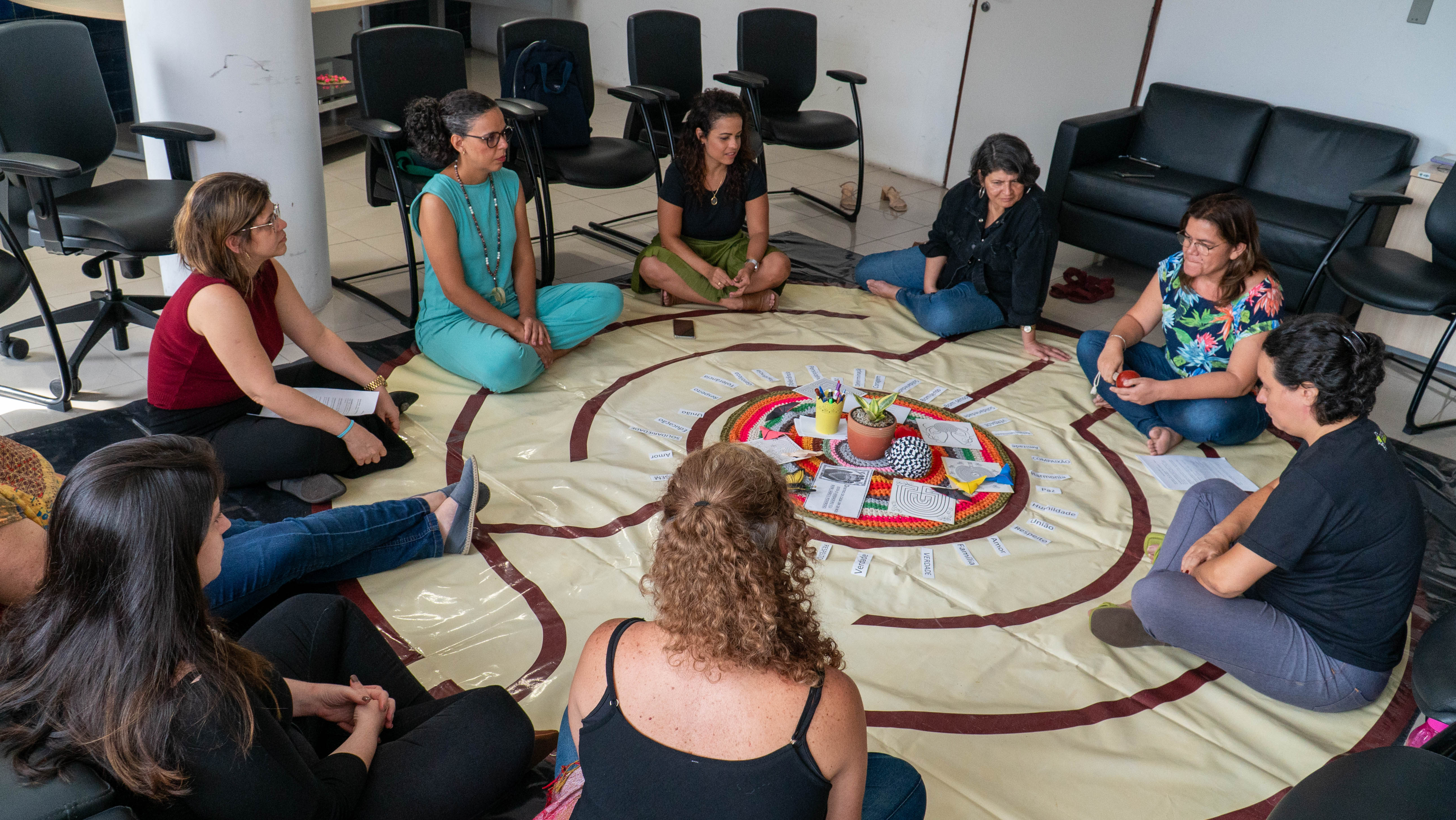 Foto mostra mulheres sentadas em círculo, no chão, em um tapete em forma de labirinto redondo
