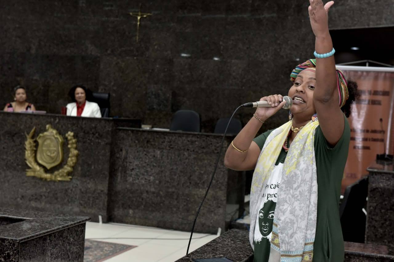 Foto mostra mulher negra falando ao microfone e outro braço levantado