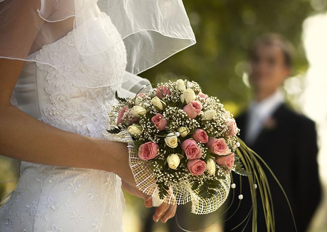 Foto mostra imagem de uma noiva segurando o buquê de flores. O noivo também está na foto.