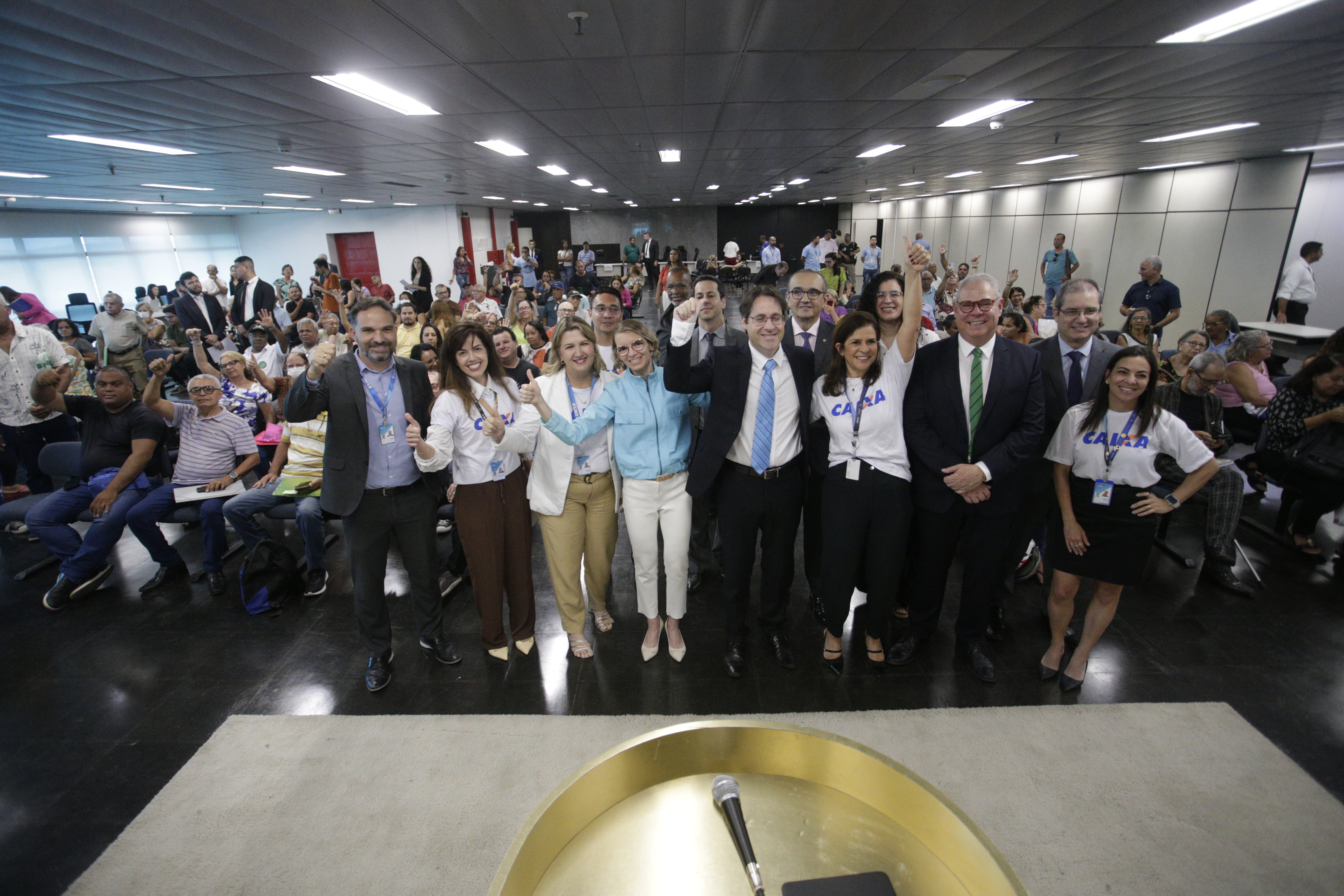 Auditório do TRF repleto de pessoas sentadas. Membros do Poder Judiciário e da Caixa Econômica em frente à câmera. Todos levantando as mãos em sinal de celebração.