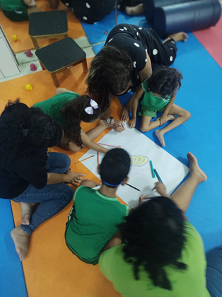 Crianças usando farda verde, sentadas em chão colorido, desenham em cartolina branca. Professoras, com uniformes escuros, também sentadas no chão, acompanham a atividade.