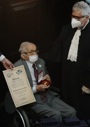 Senhor, em cadeira de rodas, recebe diploma ao lado de home de pé. Por conta da pandemia de covid-19, ambos usam máscara, em razão do período da foto, em 2022.
