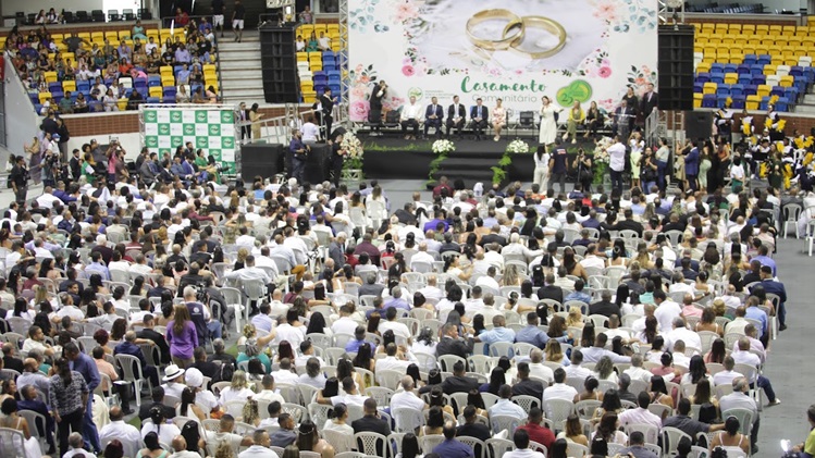 Foto mostra quadra lotada de casais sentados. Ao fundo, um palco onde há um banner enorme com a expressão "Casamento Coletivo"