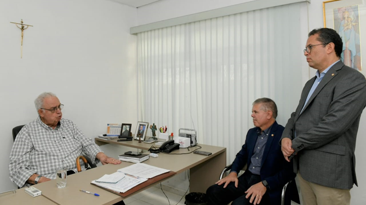 Dois homens sentados e um em pé, conversando em escritório.