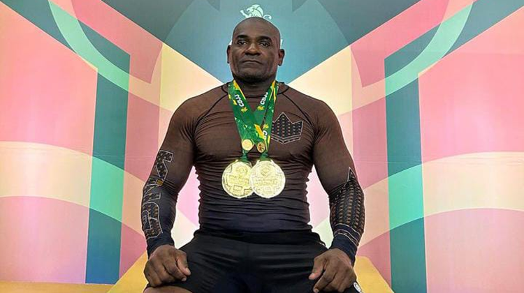 Em frente a painel colorido nas cores verde, vermelha e amarela, um homem negro e forte, usando uniforme preto, está com duas medalhas de ouro no pescoço.