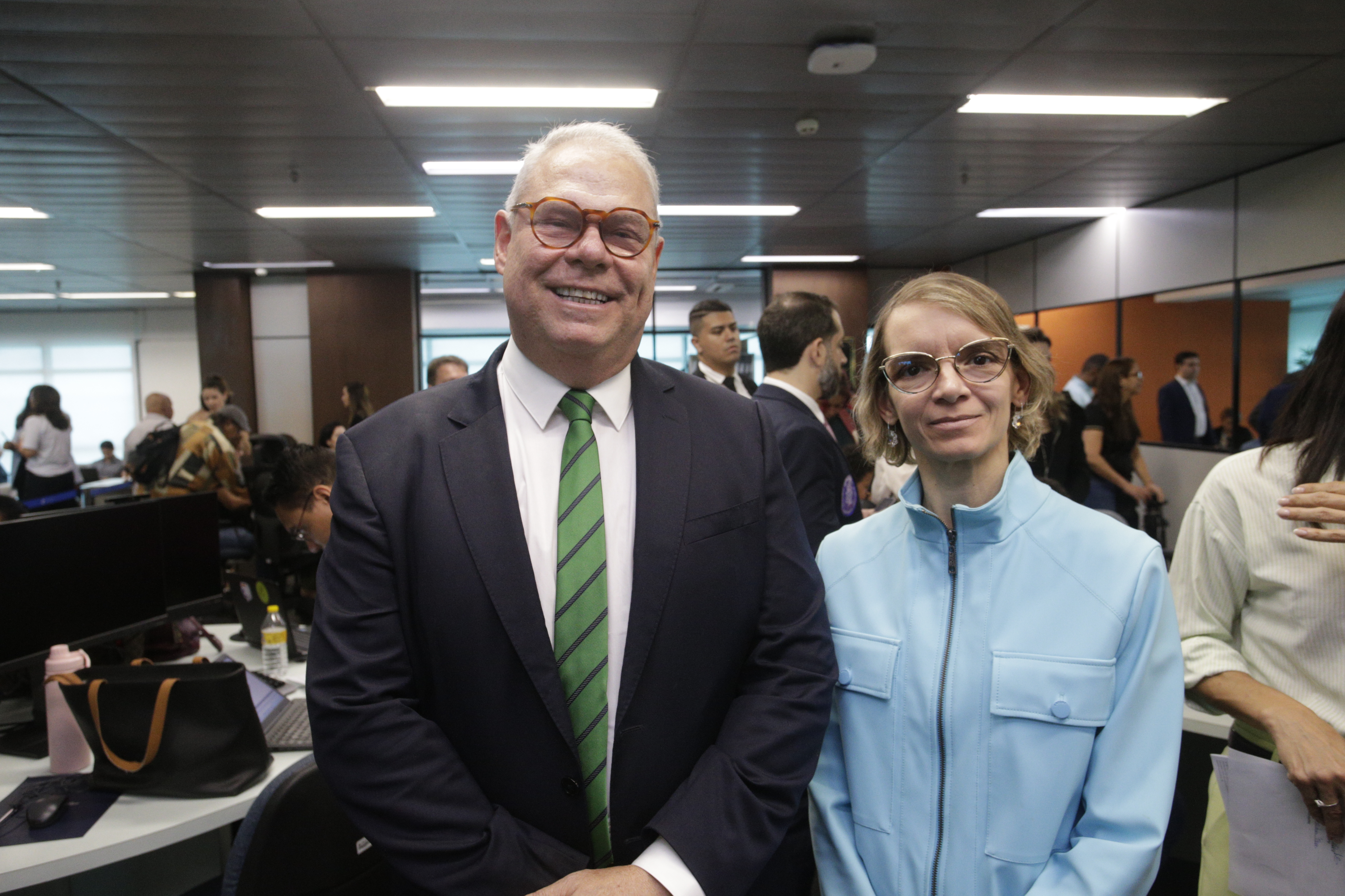 Erik Simões é um homem alto, branco, de cabelos brancos, óculos, usando um terno azul marinho e gravata verde. Joana é uma mulher baixa, branca, de cabeço curto loiro e usa óculos e trajes azuis claros