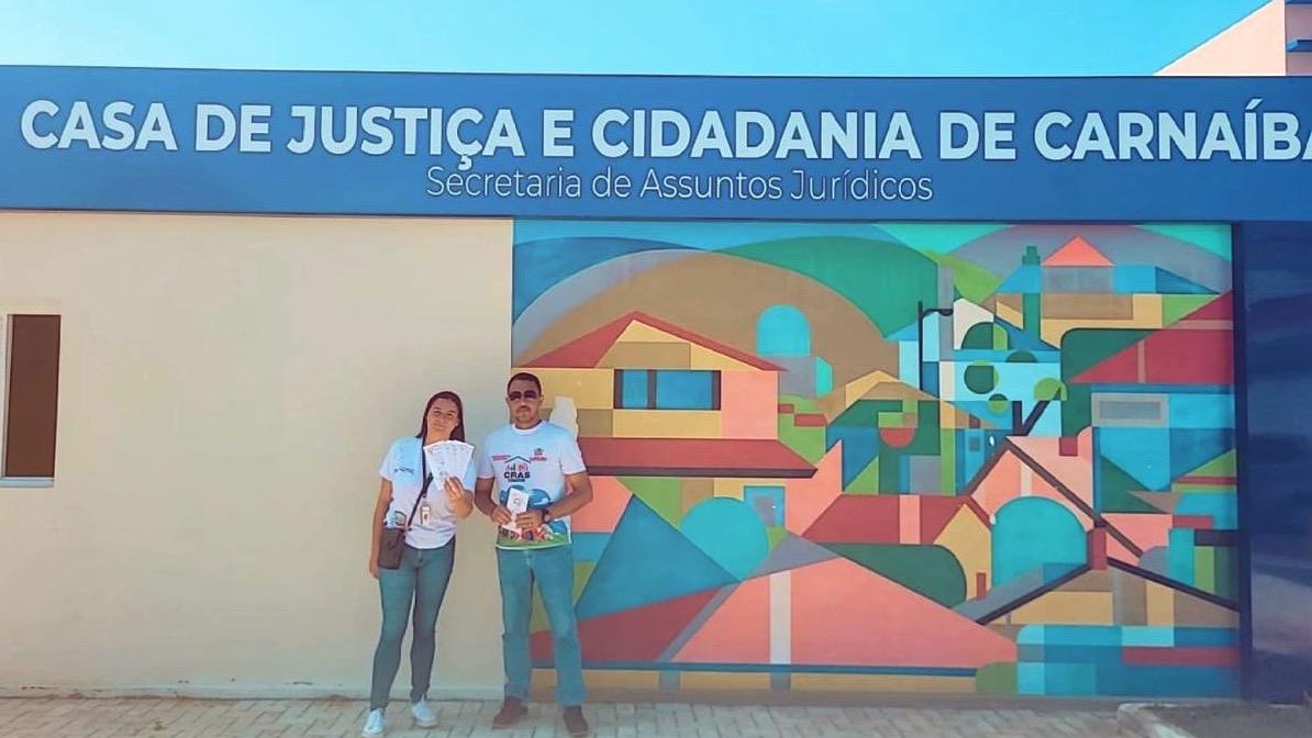 Dois servidores em frente à Casa de Justiça de Carnaíba de dia. O muro é colorido com um mosaico que lembra a arquitetura da cidade.