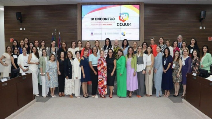 Imagem com várias magistradas e servidoras reunidas em um palco posando para foto em frente ao telão com título dos eventos juntos.