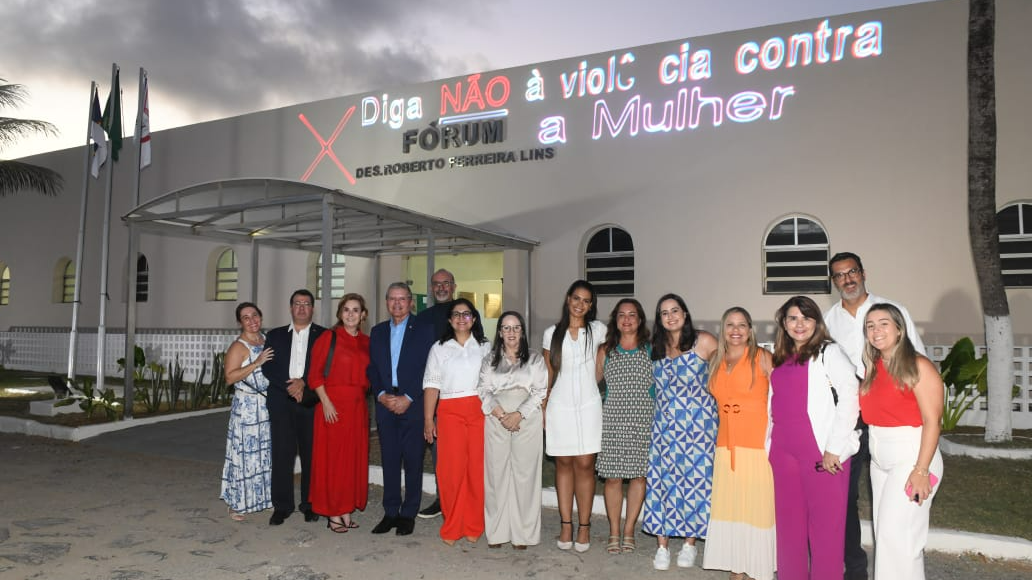 Representantes do TJPE e da administração de Fernando de Noronha na frente do Fórum no entardecer. Na parede do Fórum está projetado: "Diga NÃO à violência contra a mulher".