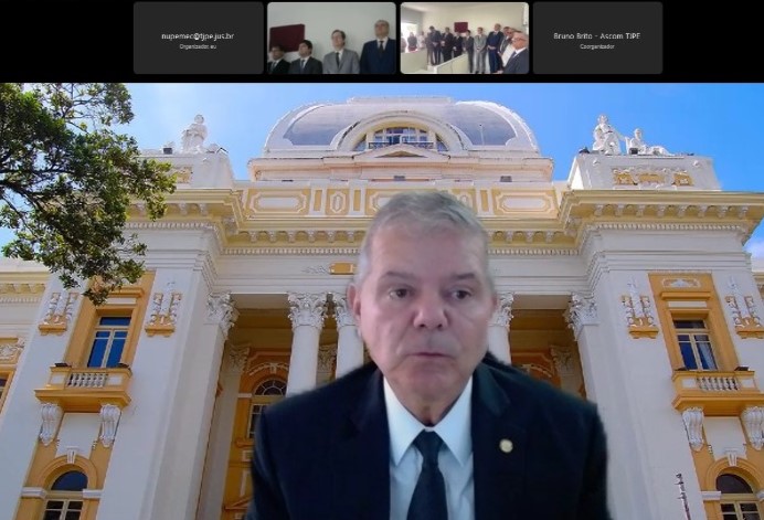 Imagem do desembargador Ricardo Paes Barreto presidindo a inauguração de Cejusc Surubim por videoconferência. O magistrado veste terno e gravata. Está no centro da imagem e ao fundo, há uma foto do Palácio da Justiça.