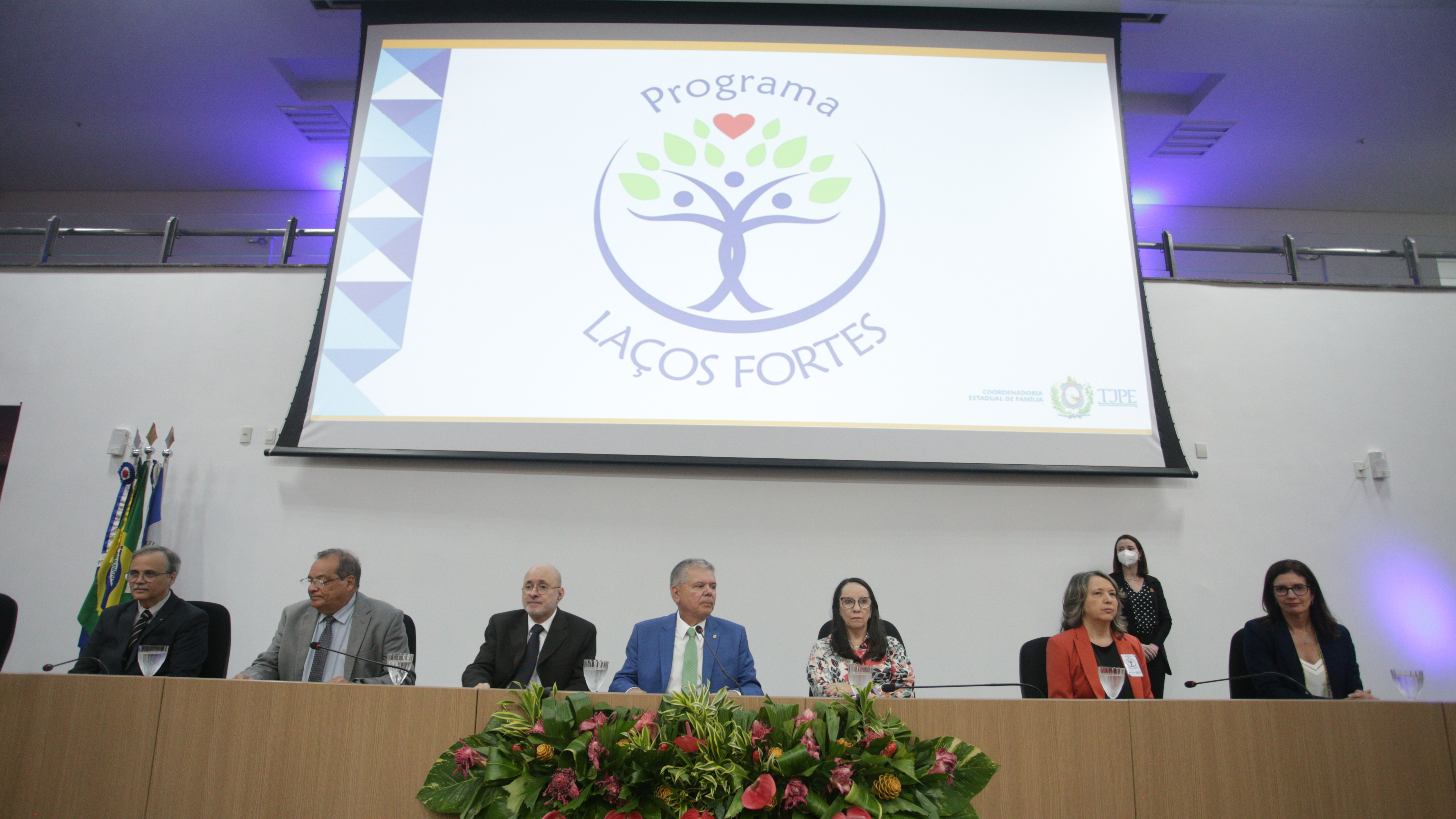 Mesa de abertura do Programa com a presença dos representantes e, ao fundo, uma projeção da logomarca do Programa: uma árvore azul com troncos trançados simbolizando três pessoas juntas com folhas verdes.