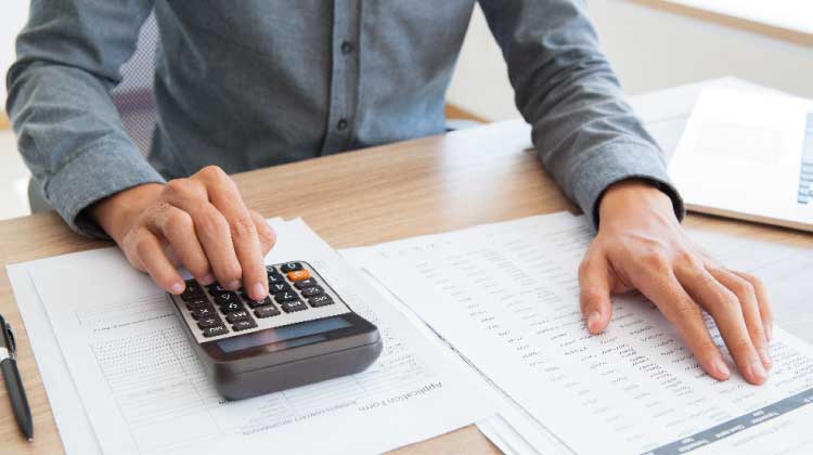 Foto de homem sentado em frente a uma mesa de madeira, usando uma calculadora e revisando números em uma lista