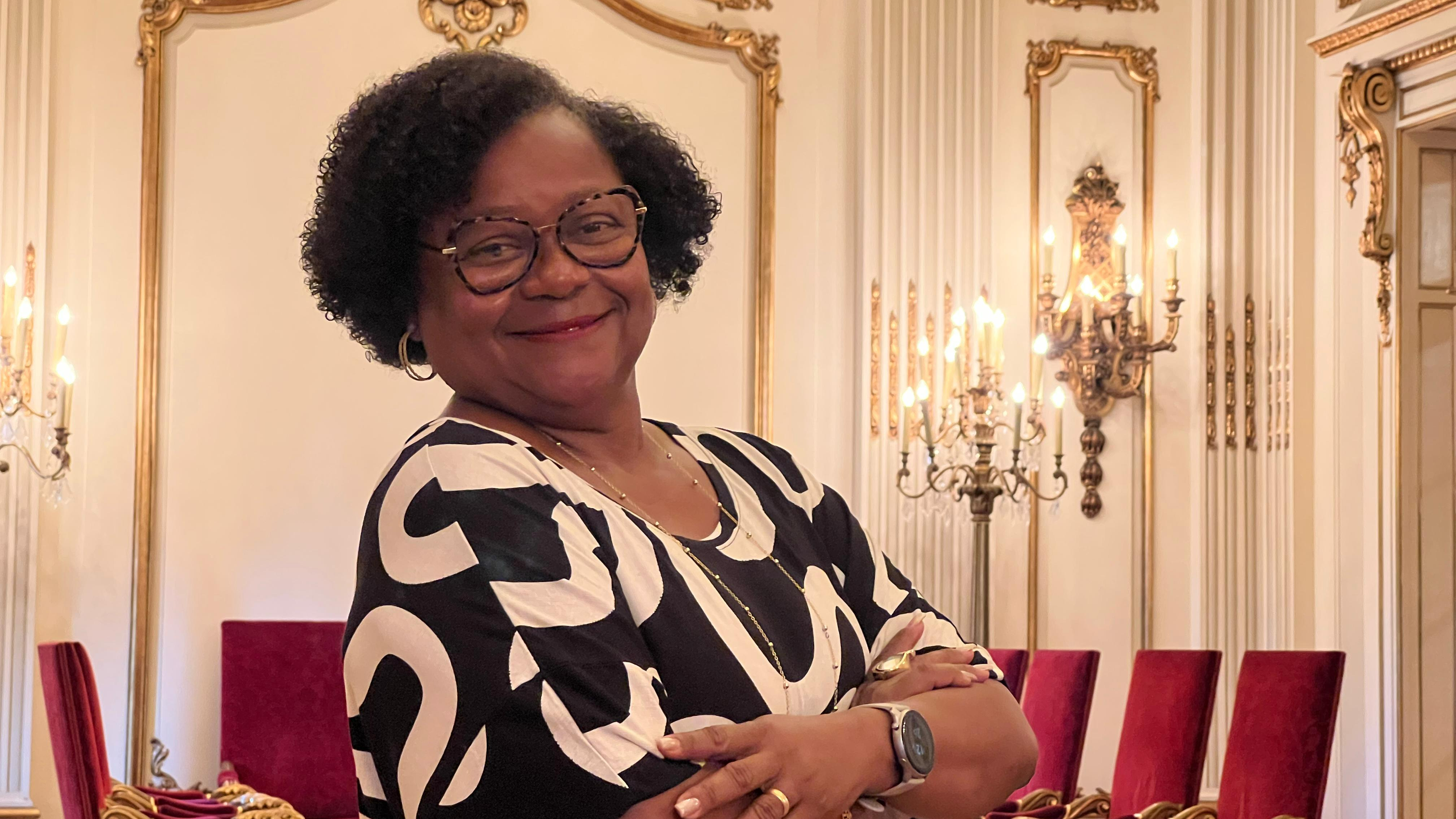 Foto de Maria da Luz no salão nobre do TJPE de braços cruzados e sorrindo para a foto. Ela é uma mulher negra, de óculos, cabelos cacheados curtos e um vestido preto com faixas curvadas brancas.