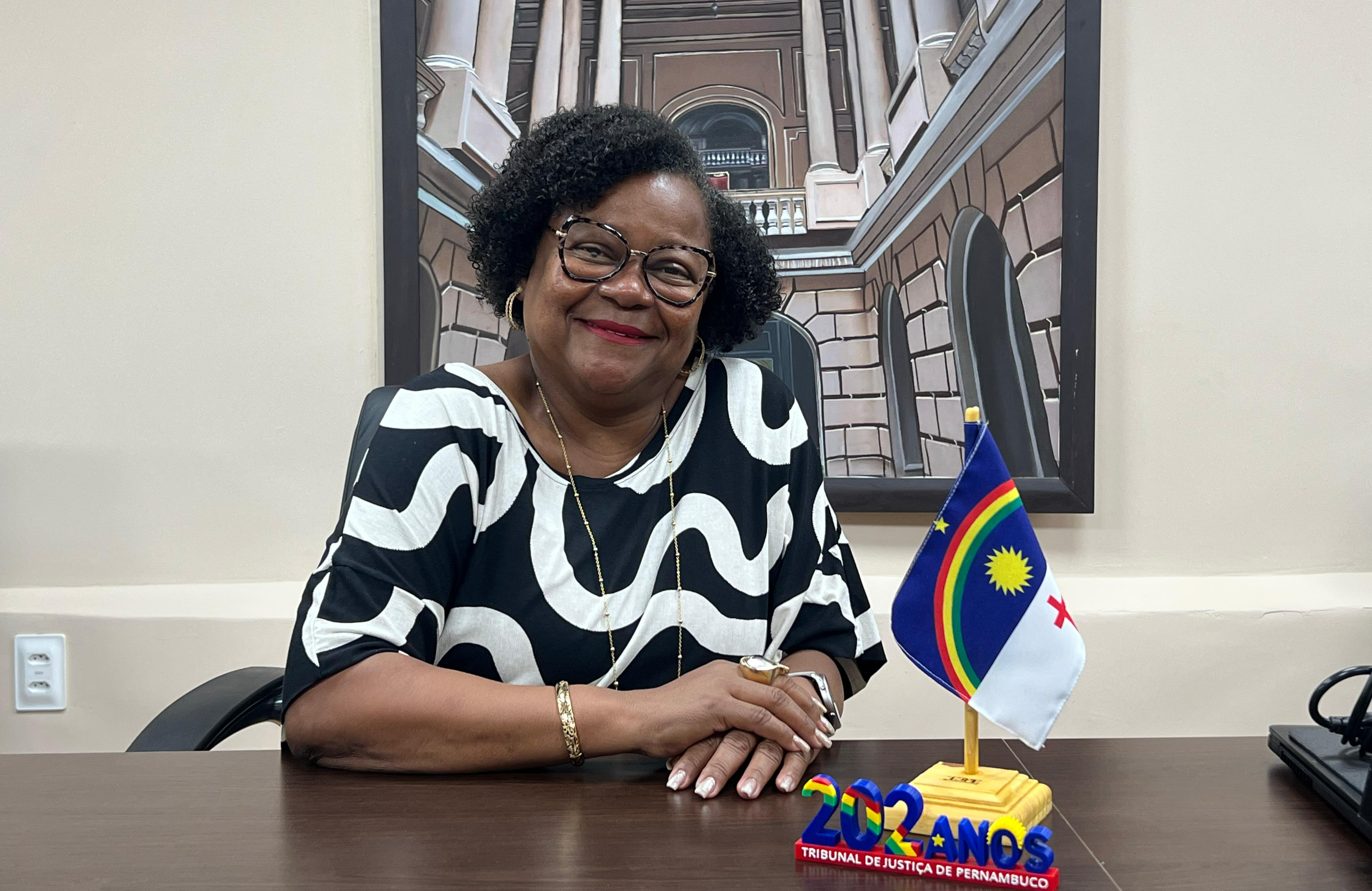 Foto de Maria da Luz na sala do Conselho de Magistratura, sentada à mesa com as mãos uma sobre a outra, sorrindo para a foto. Ela é uma mulher negra, de óculos, cabelos cacheados curtos e um vestido preto com faixas curvadas brancas. Na mesa há uma pequena bandeira de Pernambuco e uma placa de 202 anos do TJPE.