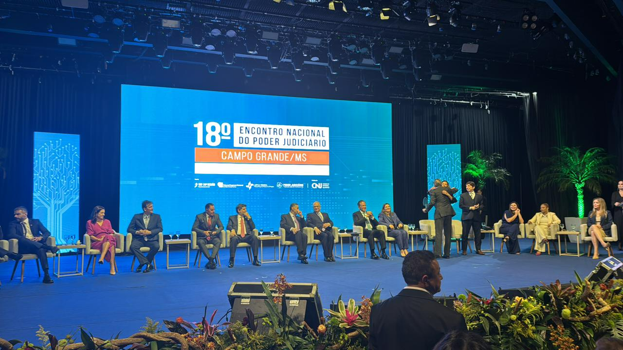 Palco com diversos membros do poder Judiciário do país sentados em semicírculo. Ao fundo, um telão de LED azul com o nome do evento.