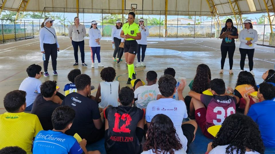 Árbitra Priscila Fernandes, uniformizada com vestes da profissão, palestrando para crianças e adolescentes em uma quadra de futsal coberta.