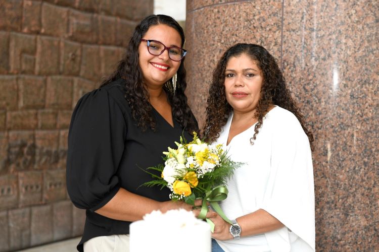 Ana Cláudia e Ana Paula são mulheres de pele parda e cabelos cacheados. Cláudia usa roupas pretas e óculos e Paula usa roupas brancas. Ambas seguram o buquê.