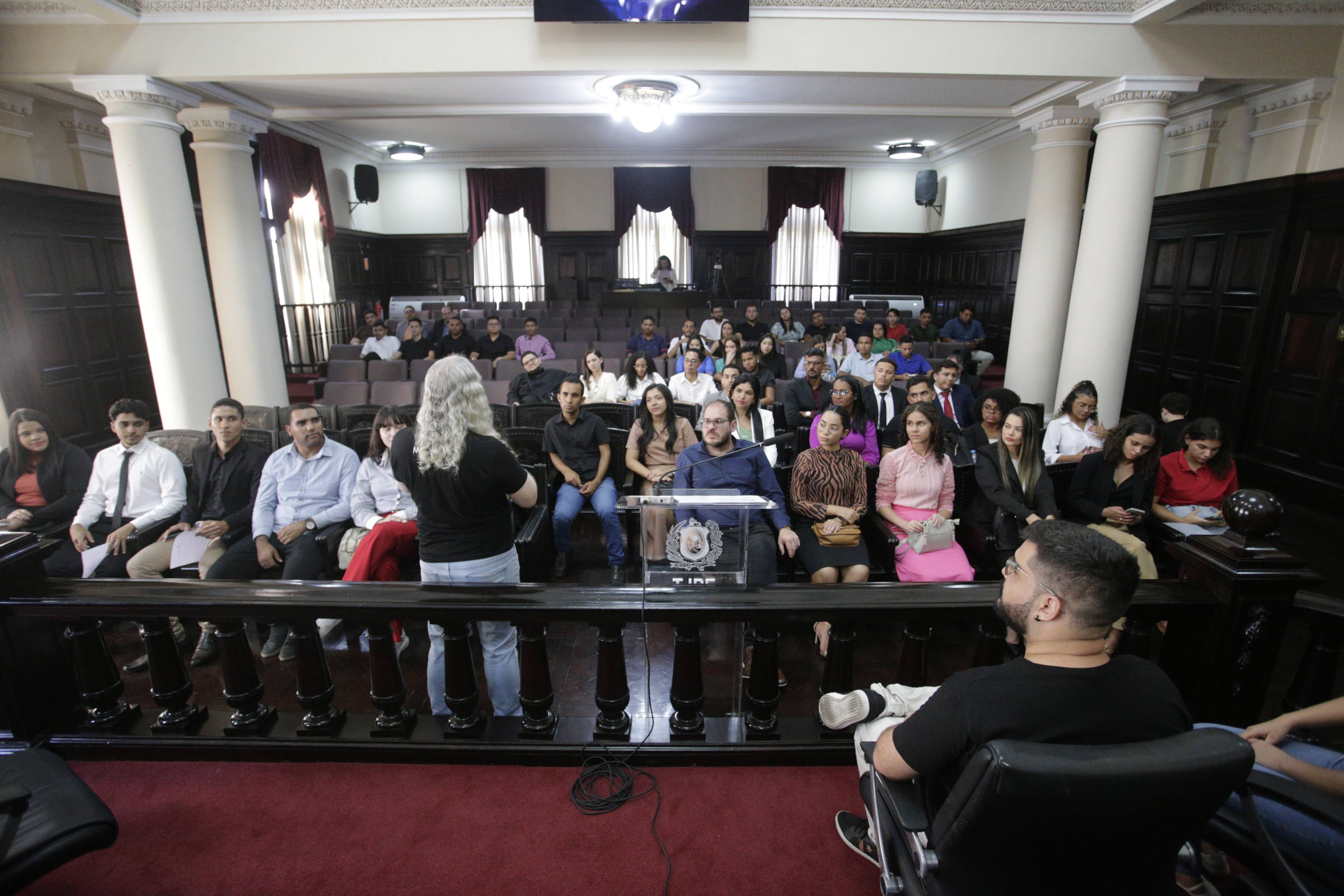 Alunos e alunas espalhados nas cadeiras do Salão Pleno do Palácio da Justiça. Os estudantes assistem a uma explicação realizada por integrante do Memorial da Justiça. 